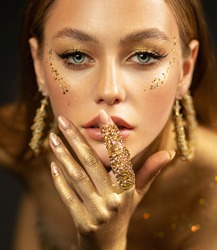 Stylish girl looks at camera. Gold ring on finger, earrings rings. Beautiful face, blue eyes, blonde hair, golden glamorous makeup, gold paint on body, skin, hands. Portrait of fashion model woman