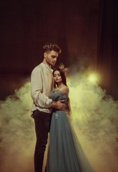 Fantasy couple hugging in dark room full white smoke. Image of lovers - king and queen. Medieval male prince in golden crown, vintage costume clothing. Girl Princess in long glamorous blue dress gown