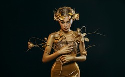Beauty fantasy woman, face in gold paint. Golden shiny skin. Fashion model girl, image goddess. Glamorous crown, wreath roses, jewellery accessories. Professional metallic makeup. hand holds a branch