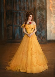 Girl beauty fantasy princess in yellow long historical, medieval silk dress holding flower red rose in her hands. Background of old gothic castle room. Fairy tale bewitched queen. Happy woman smiles.