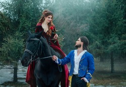 Medieval couple in love. man meets woman and gives rose in winter forest. Vintage clothing red long dress. Blue costume tailcoat caftan. Prince and princess together. Black horse. redhead hairstyle