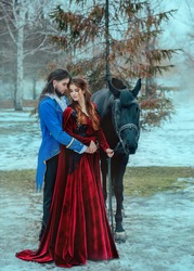 Medieval couple in love man and woman hugging in winter forest. Vintage clothing red long dress. Blue frock coat costume, tailcoat caftan. Prince and princess together. Black steed horse. Art image.