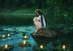 Slavic girl sits on stone on shore lake. Nymph fantasy woman hugs knees. Long black hair. Wreaths of grass, flowers float on water. Candles burning. River dusk forest green tree. Riutal of Divination.