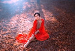 Fantasy young woman in red oriental silk dress sits on fallen orange leaves. Lady Princess in gold crown, rubies. Queen in style of Ottoman Empire. Arab beauty girl on background autumn sunny nature