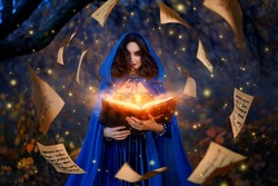 Fantasy woman witch magician in hood holds in hands magic book, bright orange light spells, wind scatters fall sheets paper page levitation. Girl sorceress. Medieval cloak blue dress magician costume
