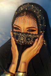 portrait close-up, young beautiful oriental woman. Beauty girl face hidden by golden veil head covered black scarf. Luxurious evening arabic style make-up, smoky green eyes. golden shadows