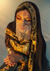portrait oriental woman sit on sand desert at sunset. Girl face hidden by gold veil head covered scarf. Evening bright arabic style make-up, smoky eyes. golden shiny black abaya traditional dress.