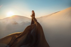 Mystery arabic woman in black long dress stands in desert long train silk fabric fly flytter in wind motion. clothes gold accessories hide face. Oriental fashion model. Sand dunes background sunset
