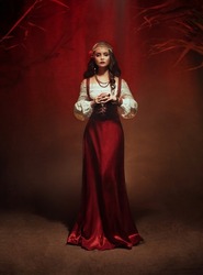  Art photo with noise. Fairy gypsy woman medieval witch stands in dark room. Long black hair, rose hairpin. Red costume vintage dress, fortune teller costume Gold jewelry . Mystical fantasy girl, 
