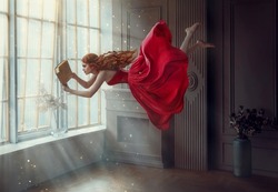 Fantasy redhead woman soars floats flies in air. Art photo levitation. Girl fairy princess reads magic book, divine light from window. Red midi dress, lon hair flutters in wind. Room classic interior