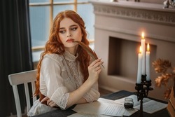 Portrait red-haired vintage woman writer holding bird feather in hands. Medieval girl sit at table writes book, letters on sheets paper with ink pen. Photo old retro style. White blouse, classic room.