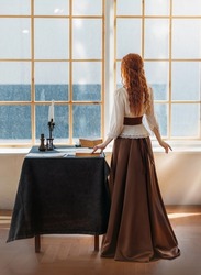 Red-haired woman in vintage dress stands looks at classic window waiting love. Clothing costume countess old style white blouse, brown long skirt. Curly red hair. Redhead girl princess back rear view.
