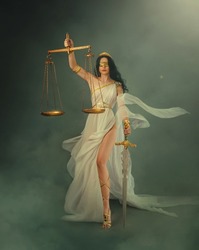 Portrait fantasy woman Greek goddess of justice Themis holding scales and sword in hands. White silk vintage dress old antique style flies waving in wind. Blind girl queen eyes blindfolded. art photo.