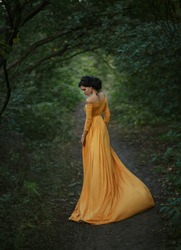 A fragile, tender girl in a yellow vintage dress . Background of a mystical arch of green trees. Artistic Photography