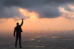 Victory and success concept! Strong confident businessman celebrating on top of skyscraper overlooking against a city sunrise background. 