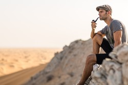 Handsome retro man sitting alone smoking a pipe on top of a rocky mountain overlooking the vast desert view.