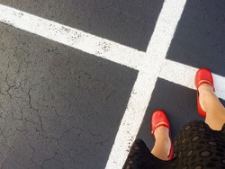A white woman wearing red  shoes and a black knee-length skirt stands to the right of white paint parking lot markings where it seems X marks the spot. Light source to the left.