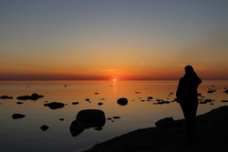 Silhouette of a woman watching sunset by calm Baltic sea with boulders, in Saulkrasti, Latvia. Sun shining from behind clouds and bright reflection on water. Selective focus