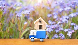 Toy blue vehicle carrying a small wooden house model against a blooming flowerbed. Creative home relocation solution concept.