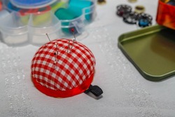 Red checkered pincushion on the white table. Needles on it. Small buttons. Tailor equipment.