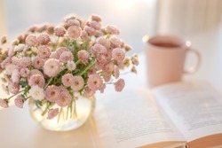 Close-up of small pink flowers bouquet in glass vase with blurred soft focused background of pink cup of tea or coffee and opened book by the window. Slow living concept.