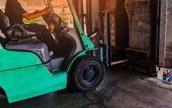 Worker driving forklift loading shipment carton boxes goods on wooden pallet at loading dock from container truck to warehouse cargo storage in freight logistics, transportation industrial, delivery