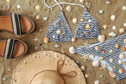 Women's summer shoes, straw hat and striped bikini set on beach sand among seashells and seafish 