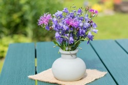On the garden table is a decorative glass vase with a bouquet of blue field bells and cornflowers.