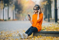 cute young girl listening music in headphones, urban style, stylish hipster teen sitting on a sidewalk on city street and choosing track on mobilephone infront of oidsity buildings, orange street