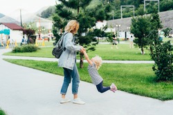 Young cute mom twirls her little adorable daughter in the city park. Emotions of a mom fooling around with a little daughter. A family walk together, happy, carefree moments. Parenting.