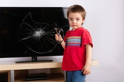 Sad little boy standing in front of a TV with broken screen holding a slingshot. Home insurance concept.