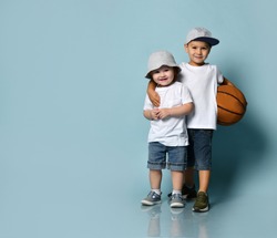 Little brunet boy hugging his toddler brother or sister. They dressed in casual clothes. Elder one holding basketball ball, smiling posing on blue background. Childhood, sport. Full length, copy space