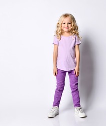 Little blonde curly girl dressed in purple blouse and pants, white sneakers. She is smiling while posing isolated on white studio background. Childhood, fashion, advertising. Full length, copy space