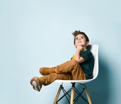 Little brunette boy in a colorful T-shirt, brown pants and sneakers. The boy looks up, propping his chin with his fist, sitting on a white chair on a blue studio background. Childhood, fashion.