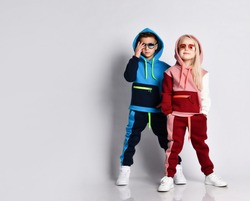 Little kids, boy and girl, in sunglasses and hoods, colorful tracksuits, sneakers. They posing isolated on white studio background. Childhood, fashion, advertising and sport. Full length, copy space