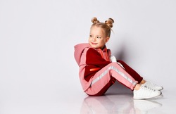 Little blonde girl with buns hairstyle, in colorful tracksuit, sneakers. She laughing, sitting on floor, posing isolated on white. Childhood, fashion, advertising and sport. Close up, copy space
