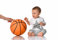 Cute baby boy in casual jumpsuit sitting on floor. Mother hand passing basketball ball to adorable toddler child. Sport game, activity and leisure. Studio portrait on white wall background