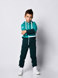 Little brunette kid in colorful tracksuit and sneakers on a gray background. Little brunette boy folded his arms and posed looking at the camera. Full length