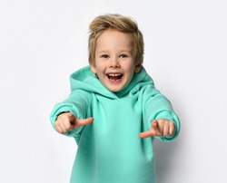 Close up portrait of a joyful active boy pointing his fingers at you. Happy cute blond boy in a warm turquoise hoodie on a gray background. Childhood, style and sportswear concept.