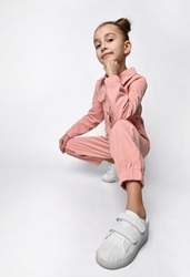 Portrait of a cute girl in a casual cotton jumpsuit with pockets, squatting with one leg in a white sneaker forward, stylish hairstyle.