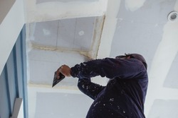 A man applies putty to a ceiling of an almost complete home. Wearing clothes full of paint splotches. Shot at the garage area.
