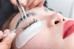 A beautician prepares to insert an individual lash into the eyelash line of an asian woman during an extension procedure.