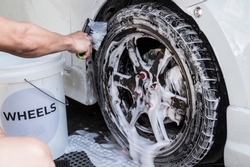 A man use a large brush to scrub clean a car's front tire and loosen dirt and grime in the grooves. At a carwash or auto detailing shop.