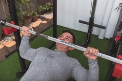 A fit asian guy performs a set of bodyweight inverted rows. Gripping on a steel bar on a squat rack. Training back at his home gym.