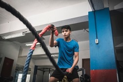 A fit young asian man working out vigorously with battle ropes. Alternating single arm waves. Whole body workout, conditioning and cardio at the gym.