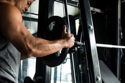 Gritty scene of a man adding heavy barbell plates on a smith machine at the gym. Focus on rubberized black plates. Gym or Fitness club setting.