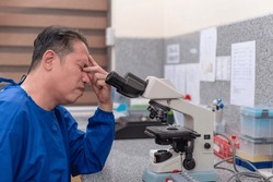 An asian male laboratory technician or microbiologist in his 40s feeling exhausted and burned out while at work. Unhealthy levels of stress or experiencing eye strain. Wearing a blue scrub suit.