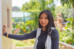 A lovely lady in a cardigan and navy blue long sleeves top is looking directly at the camera with a straight face while her hand is leaning on a wall. Plants and trees in the background.