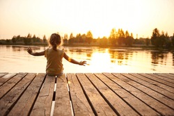 Silhouette of a child sitting on the wooden pier and enjoying heat summer evening at the lake at sunset. Heat summer, summer evenings in nature, spending time in the fresh air