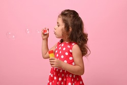 Side portrait of a cheerful beautiful baby girl blowing soap bubbles, isolated over pink background with space for text . Gorgeous child playing with soap bubbles. Summer children leisures concept